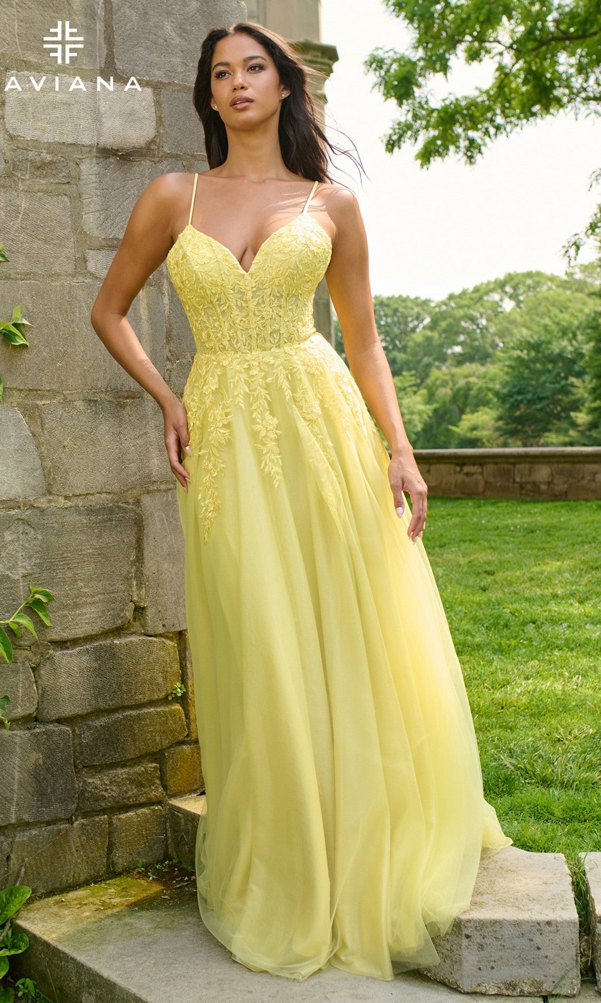 A woman stands outdoors by a stone wall, wearing the Faviana 11438 long prom dress with thin straps, floral details, and an elegant yellow a-line tulle skirt.